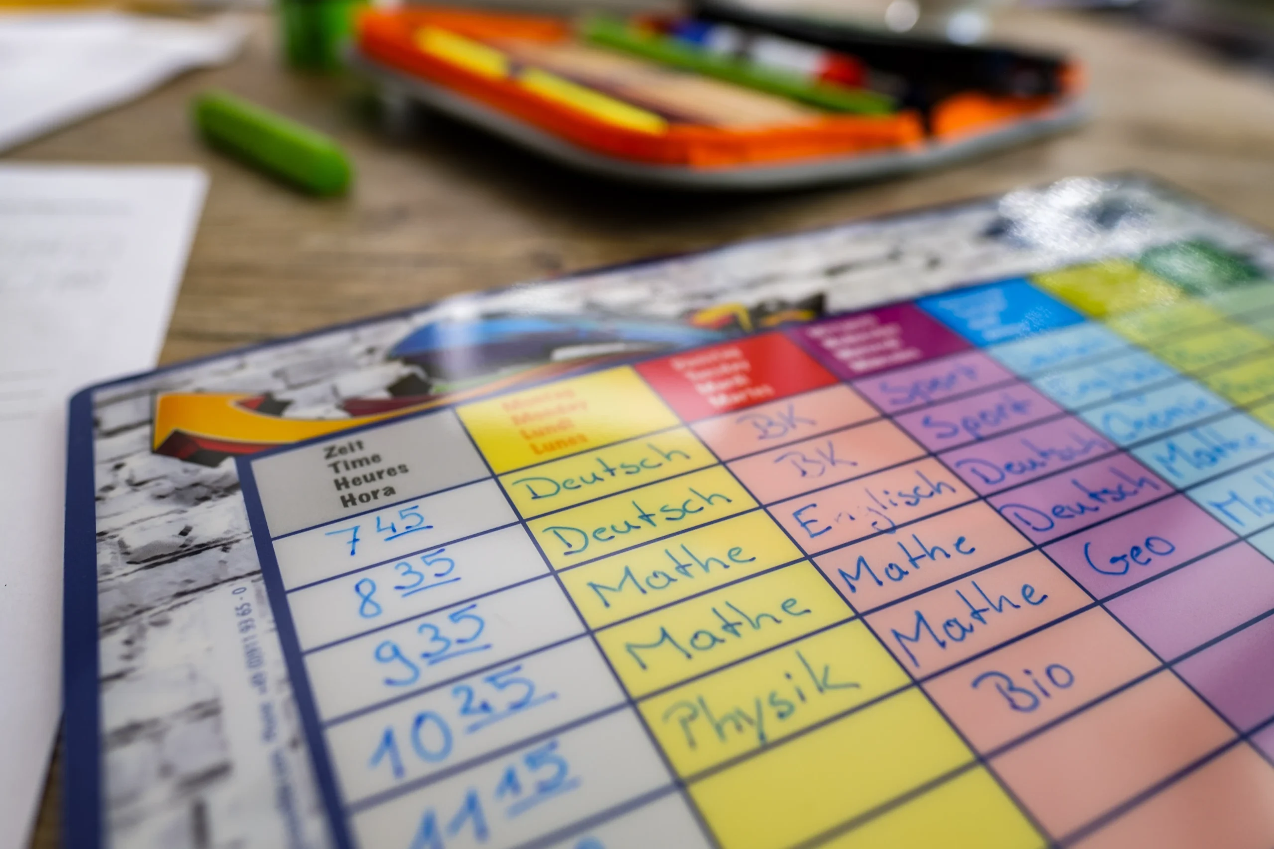 Nahaufnahme eines bunten Schulstundenplans mit Fächern wie Deutsch und Mathe. Im Hintergrund liegen ein Federmäppchen und Stifte verschwommen auf dem Tisch.
