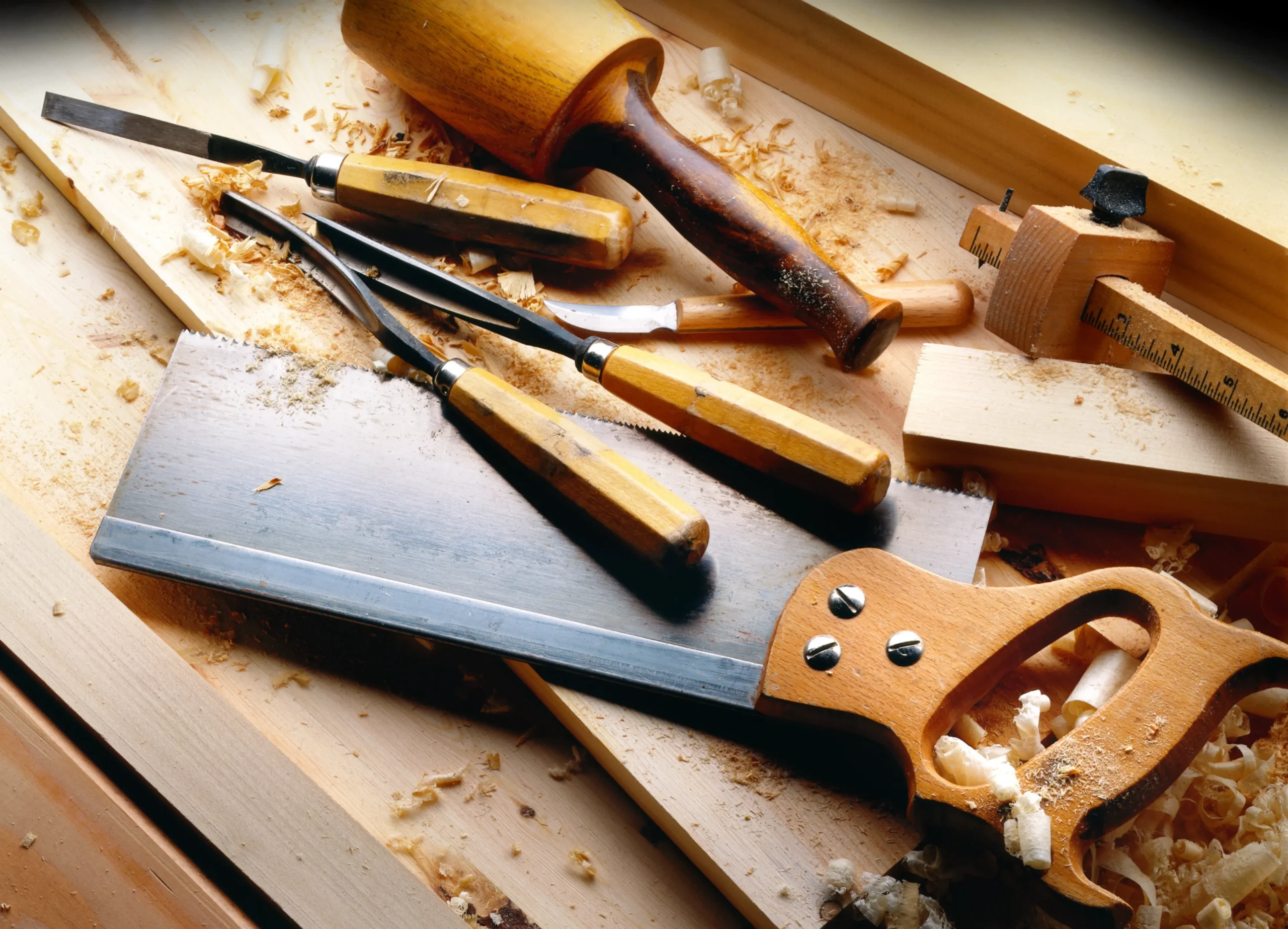 Klassische Schreinerwerkzeuge im Set, darunter eine Handsäge mit Holzgriff, Schnitzmesser, Stechbeitel und ein Holzschlägel, verteilt auf einer Hobelbank inmitten von feinen Holzspänen.