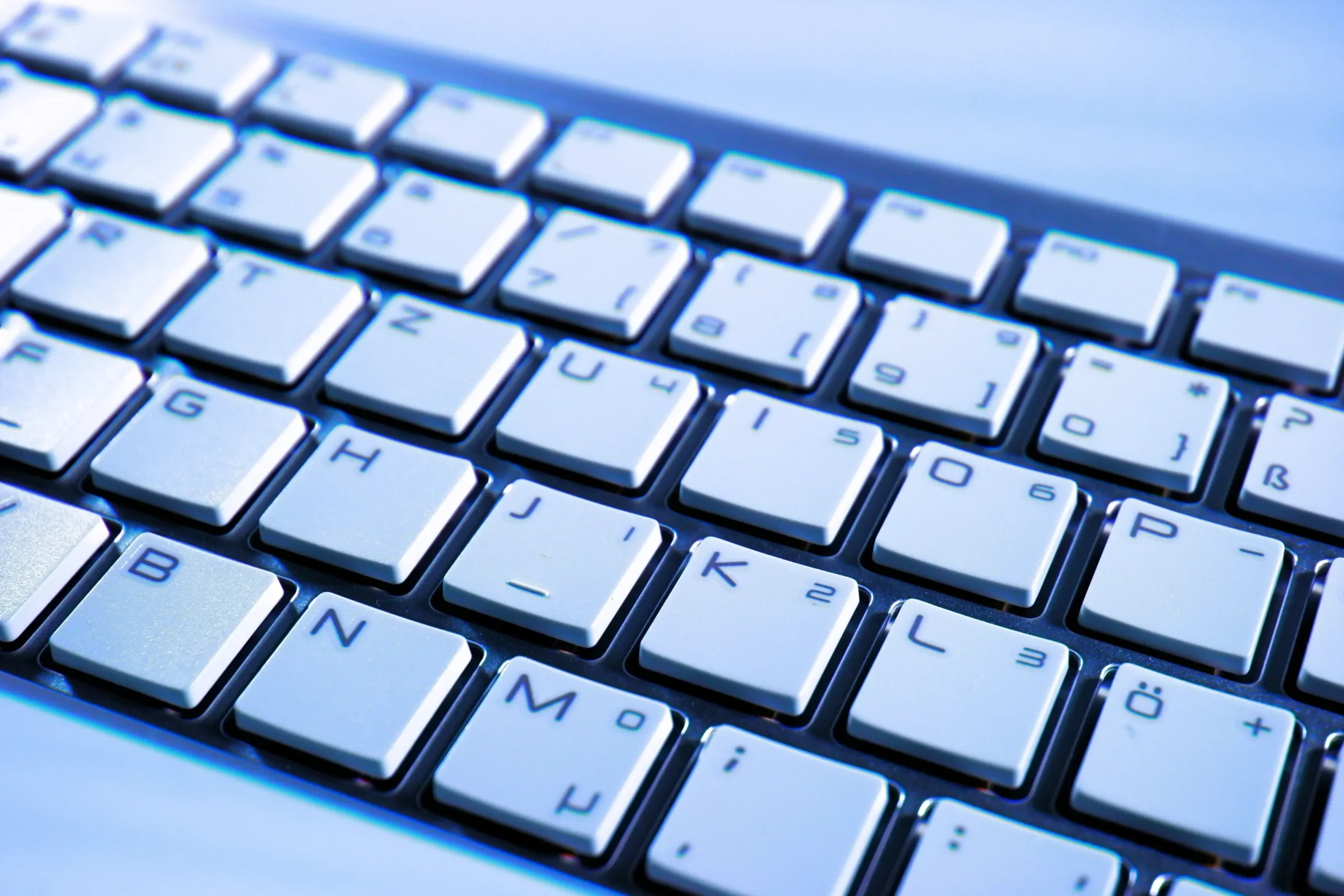 Schräge Nahaufnahme einer modernen PC-Tastatur mit flachen weißen Tasten und deutschem Layout, die kühl blau ausgeleuchtet ist.