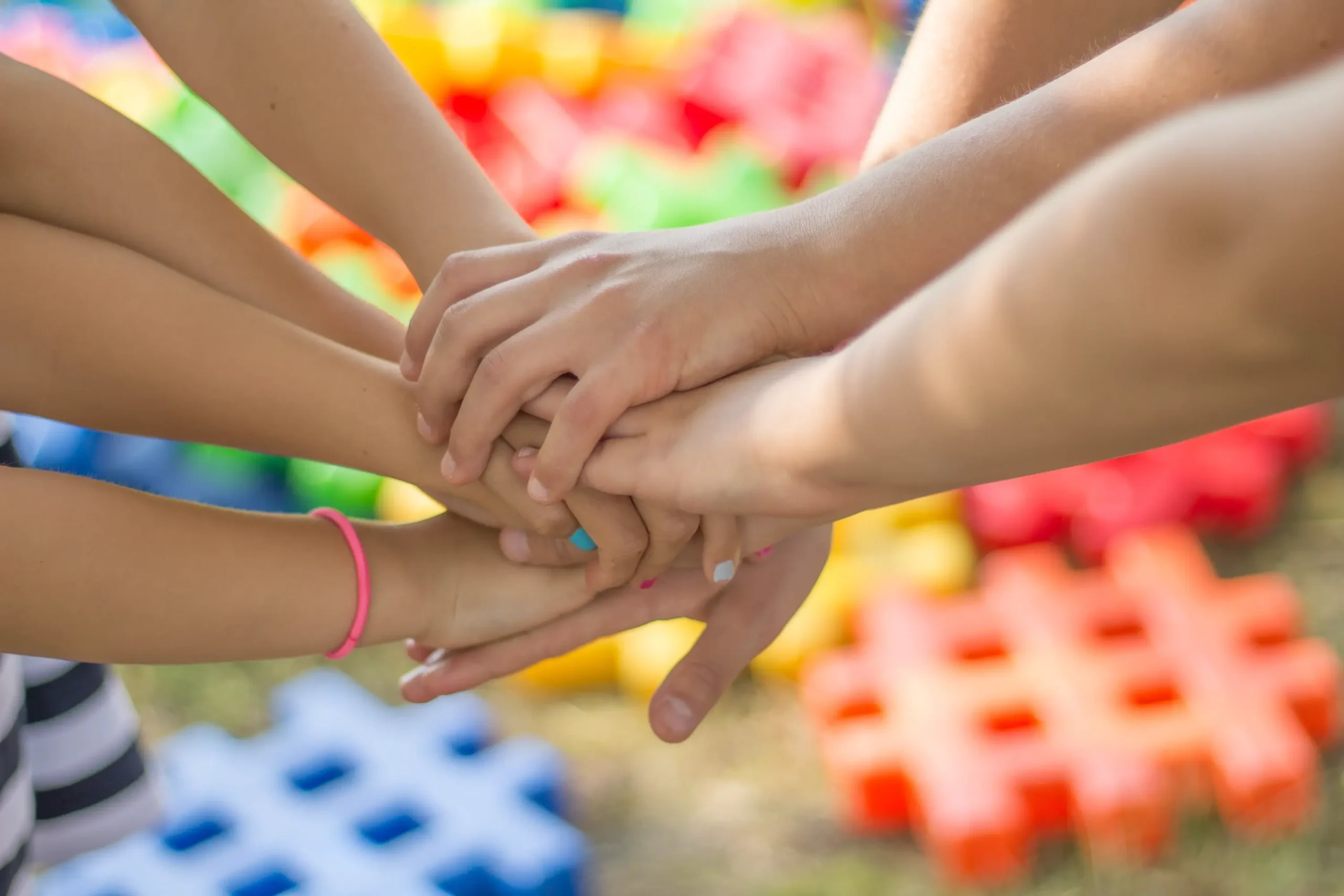 Nahaufnahme von mehreren Kinderhänden, die in der Mitte aufeinandergestapelt sind. Im unscharfen Hintergrund liegen bunte Plastikbausteine auf dem Rasen.