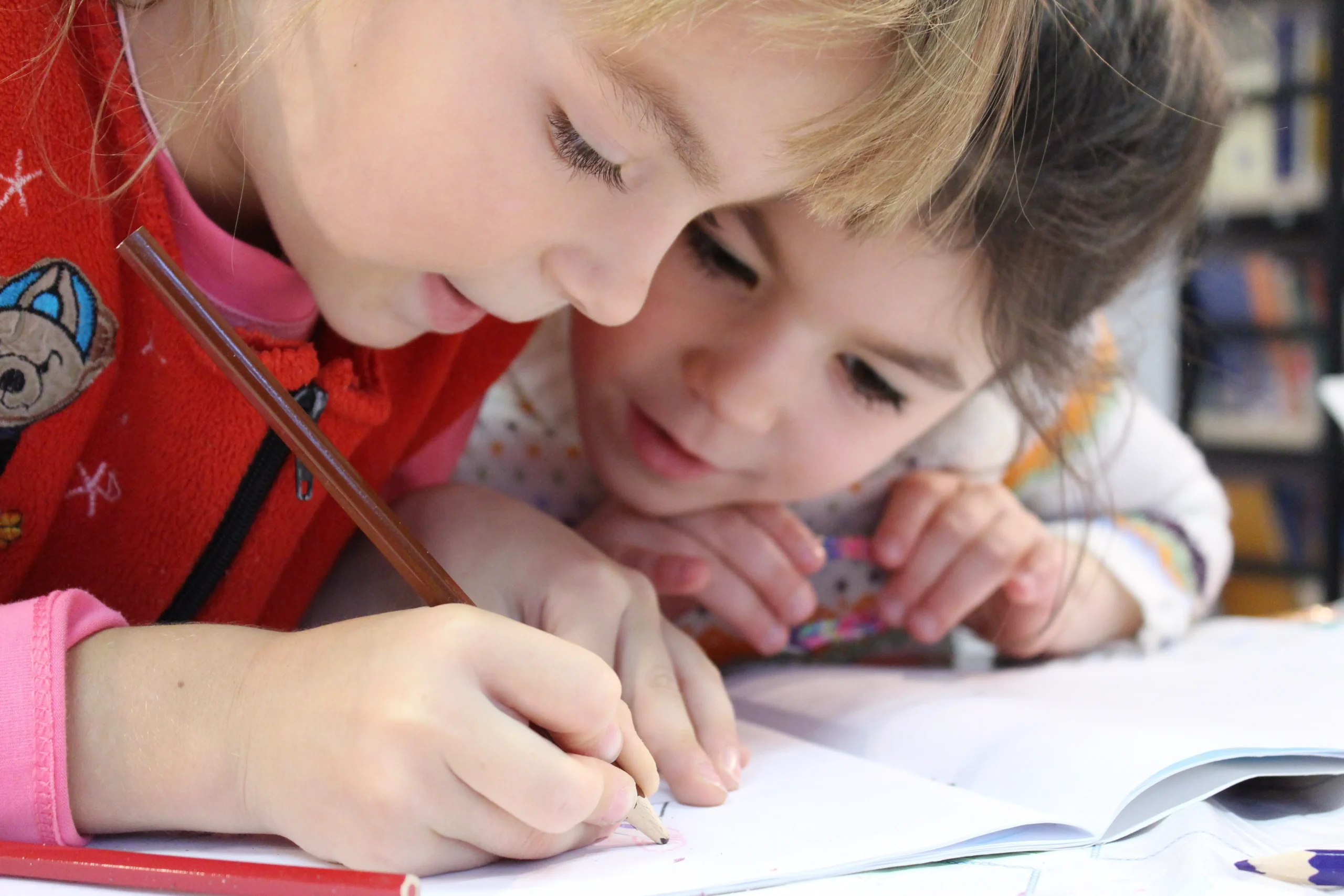Nahaufnahme von zwei Kindern, bei der ein Mädchen mit einem Stift in ein Heft zeichnet und ein anderes Kind ihr dabei über die Schulter schaut.