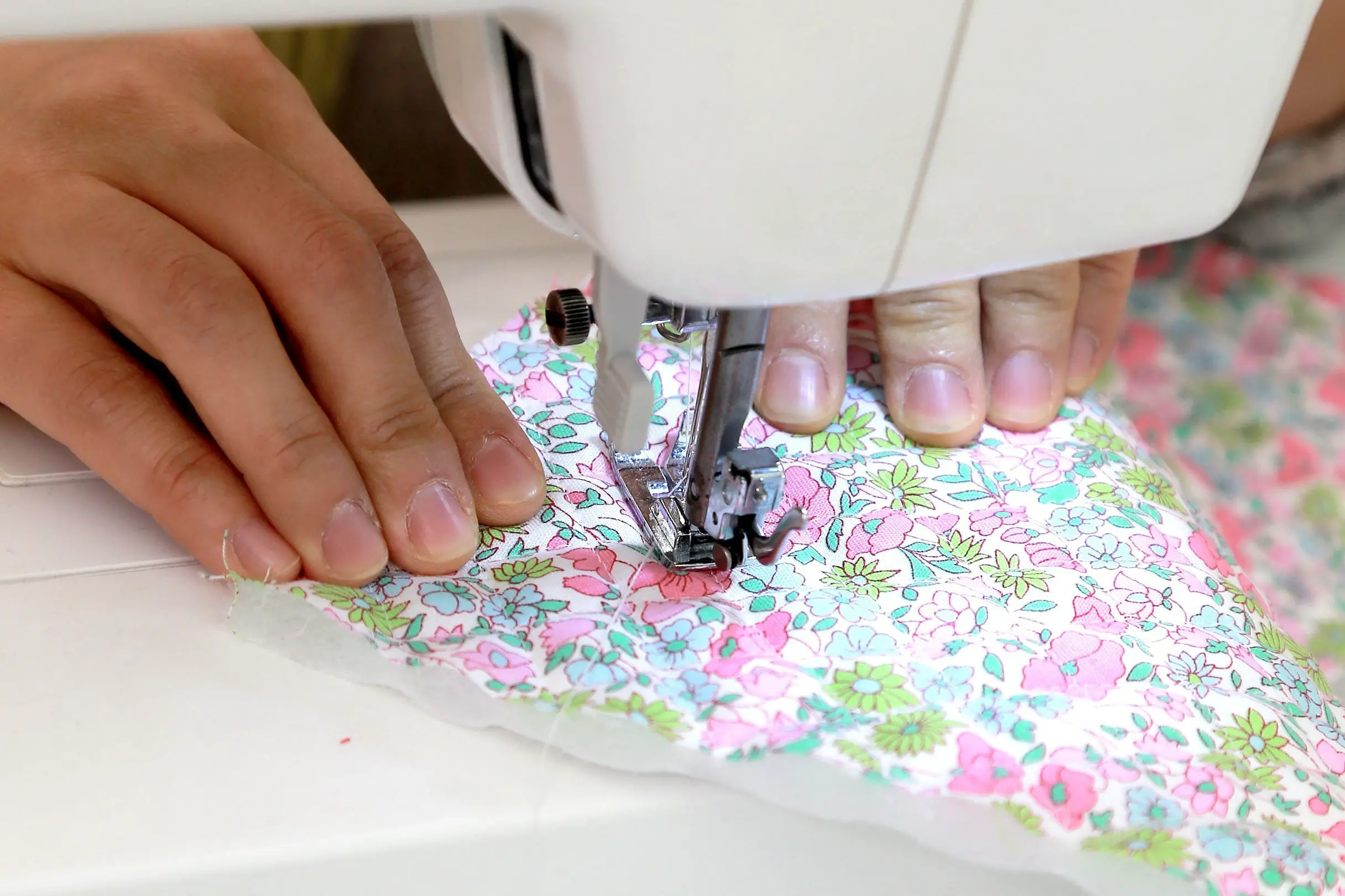 Zwei Hände führen ein Stück Stoff mit rosa und grünen Blumenmustern unter dem Nähfuß und der Nadel einer weißen elektrischen Nähmaschine.