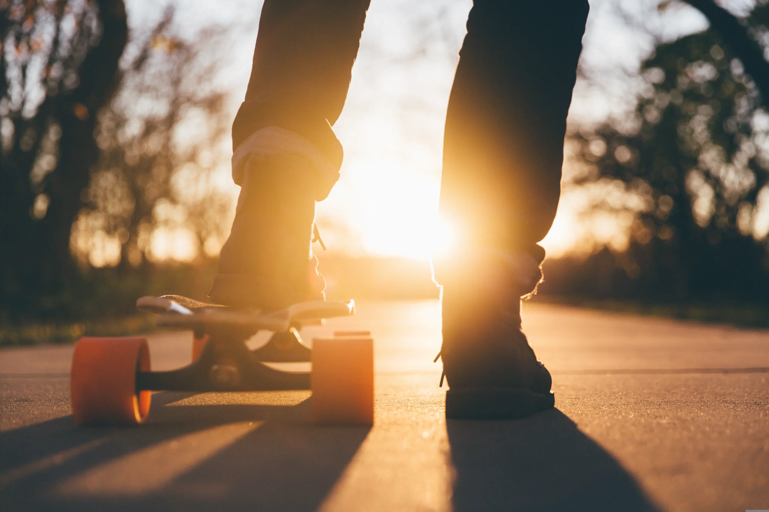Nahaufnahme von hinten auf die Beine einer Person, die mit dunklen Schuhen auf einem Cruiser-Skateboard mit orangefarbenen Rollen steht. Die tiefstehende Sonne wirft ein warmes Licht auf den asphaltierten Weg.