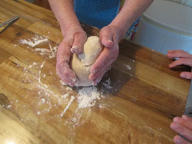 Nahaufnahme von den Händen einer älteren Person beim Kneten von Brotteig auf einem Holztisch mit Mehl. Am rechten Bildrand schaut ein Kind zu.